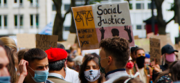 A crowd at a rally, with a sign in the background reading "Social Justice"