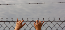 A pair of hands, reaching up to climb over a barbed wire fence, with gloomy grey skies in the background